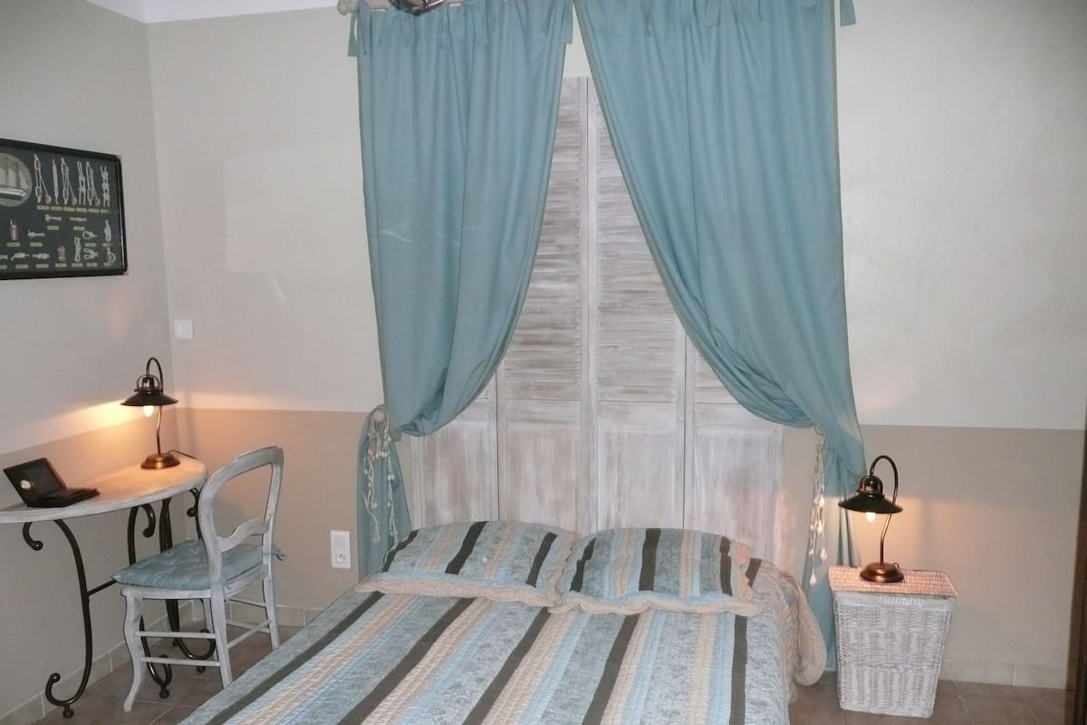 A bedroom with a bed, a desk, a chair, and a small table with a lamp.