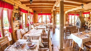 A rustic wooden interior with tables set for dining, a stone fireplace, and red curtains. - Bungalows Drevna (Novi Sad)