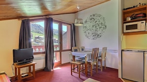 A cozy room with a TV, dining table, chairs, a small kitchen area, and a large window with curtains. - South-facing apartment at slopes, 5 people, balcony (Plagne Soleil)