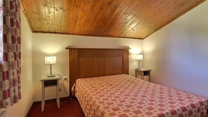 A bedroom with a wooden ceiling, a bed with a patterned bedspread, two bedside tables with lamps, and a curtain on the left. - South-facing apartment at slopes, 5 people, balcony (Plagne Soleil)