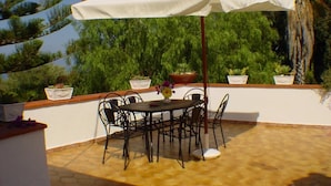 Un patio al aire libre con una mesa y sillas bajo un paraguas blanco, rodeado de plantas en macetas y una palmera.