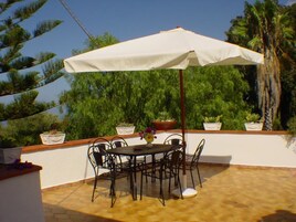 Un patio al aire libre con una mesa y sillas bajo un paraguas blanco, rodeado de plantas en macetas y una palmera.
