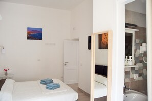 Una habitación de hotel con cama, un baño con lavamanos y espejo, una puerta y un cuadro de un paisaje colgado en la pared.