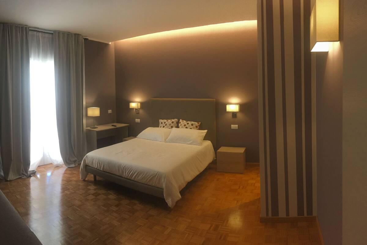 A hotel room with a bed, nightstands, a sofa, and a window with curtains.