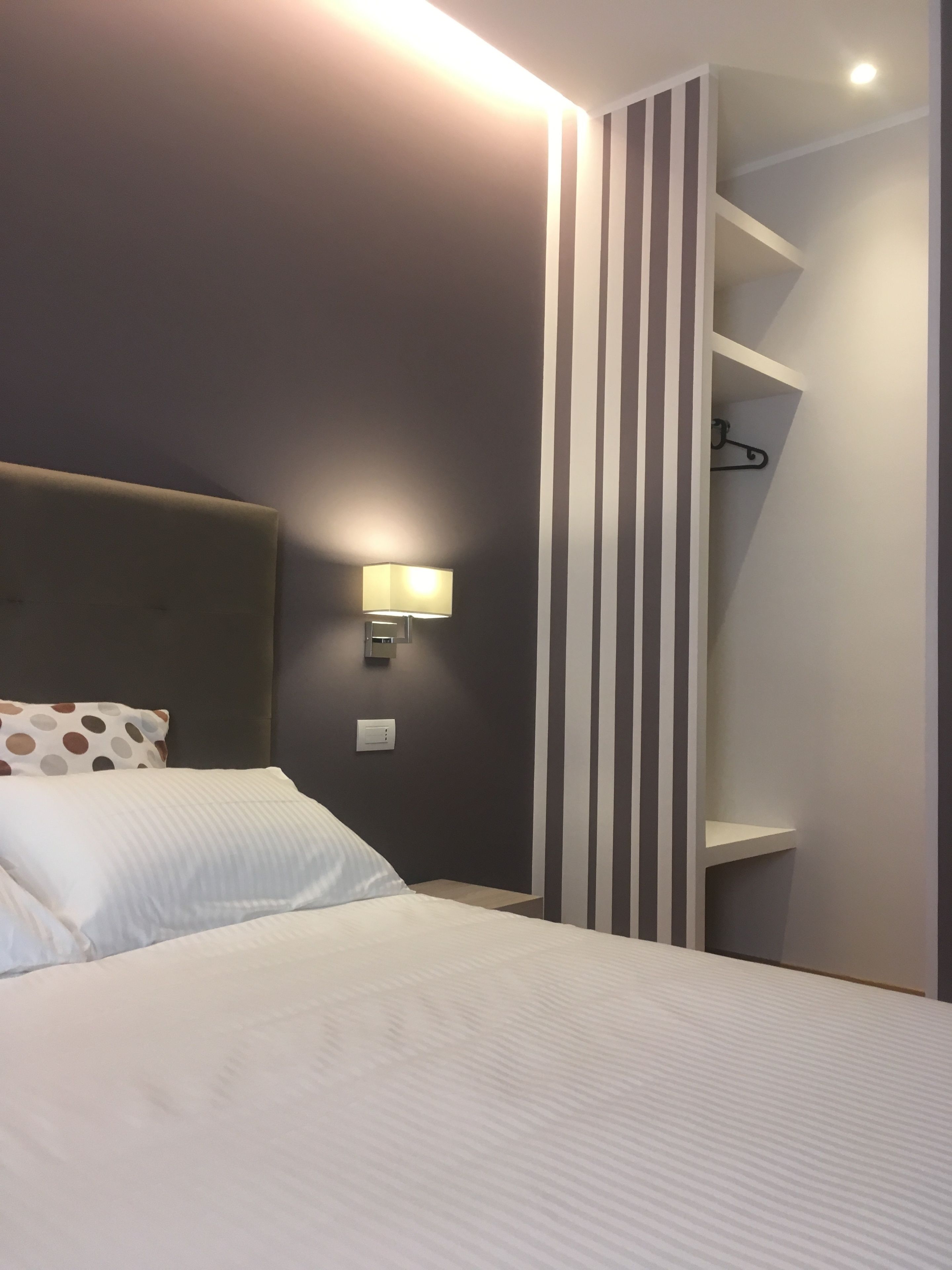 Una cama bien hecha con sábanas blancas, cabecera y una lámpara de pared.