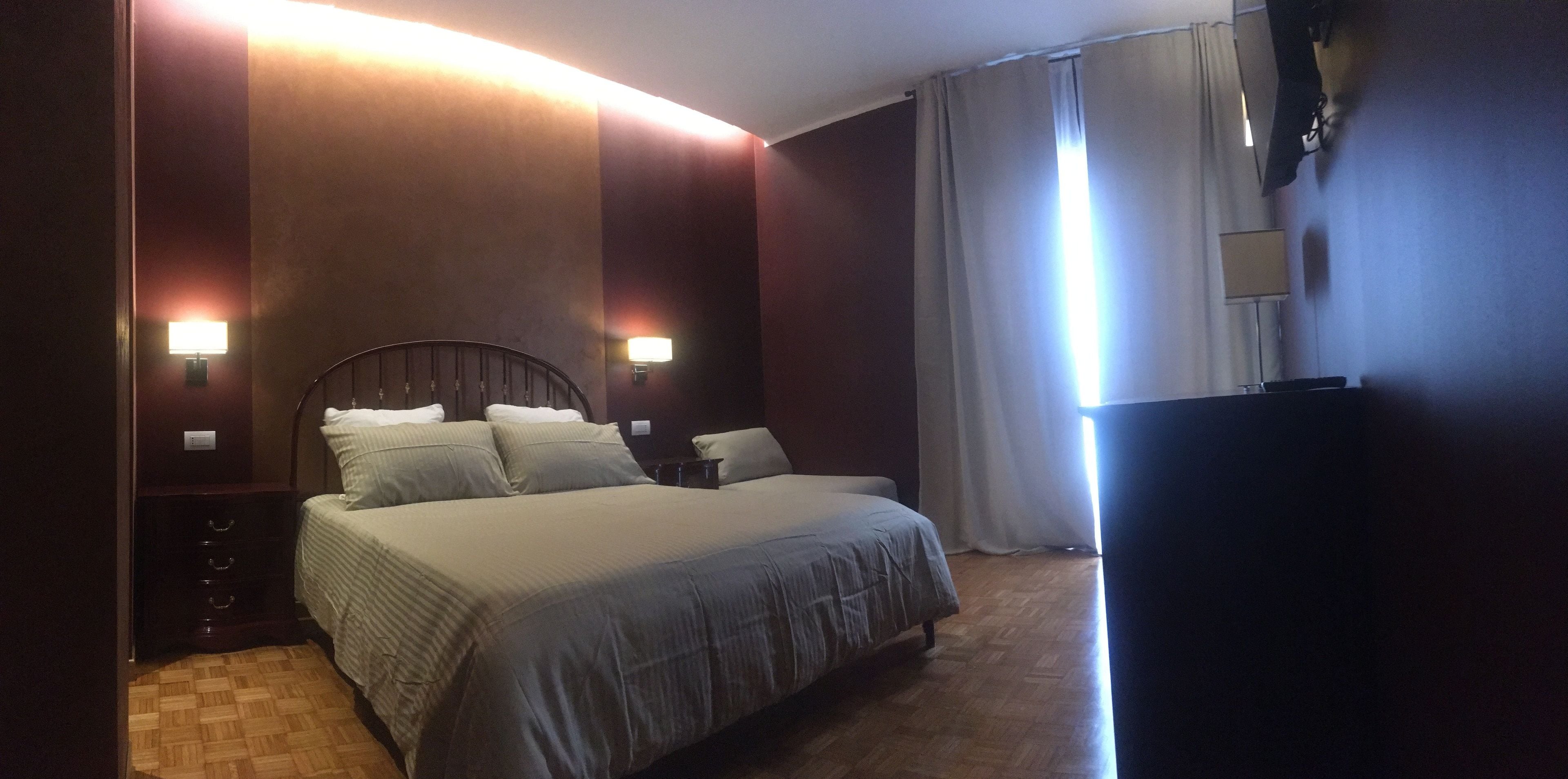 A hotel room with a large bed, two wall-mounted lamps, a wooden floor, and a TV on the right side.