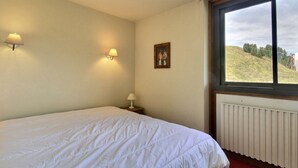 A bedroom with a bed, two wall-mounted lamps, a framed picture, and a window with a view of a hill. - Apartment 6 pax, Mont Blanc view, near slopes, parking (La Plagne)