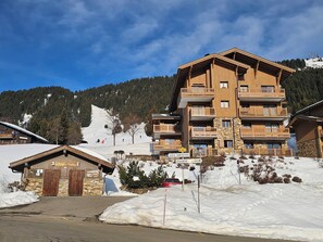 Un immeuble en bois à plusieurs étages, doté de balcons, avec une piste de ski en arrière-plan et une enseigne indiquant « Les Saisies » et « Villard X Coron ».