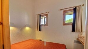 Una habitación pequeña con una cama individual, colcha roja, dos ventanas con cortinas, una lámpara de pared y un calefactor pequeño.