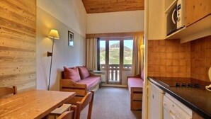 Una habitación acogedora con techo de madera, un sofá, una mesa de comedor con sillas, una zona de cocina con lavavajillas y vistas a través de una puerta.