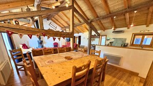 A rustic dining area with a wooden table and chairs, a kitchen in the background, and a stone fireplace.