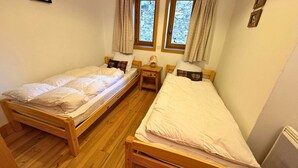 A room with two wooden beds, wooden floors, a small bedside table with a lamp, and a window with curtains.