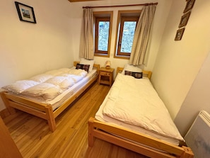 Una habitación con dos camas de madera, piso de madera, una mesita de noche con lámpara y una ventana con cortinas.