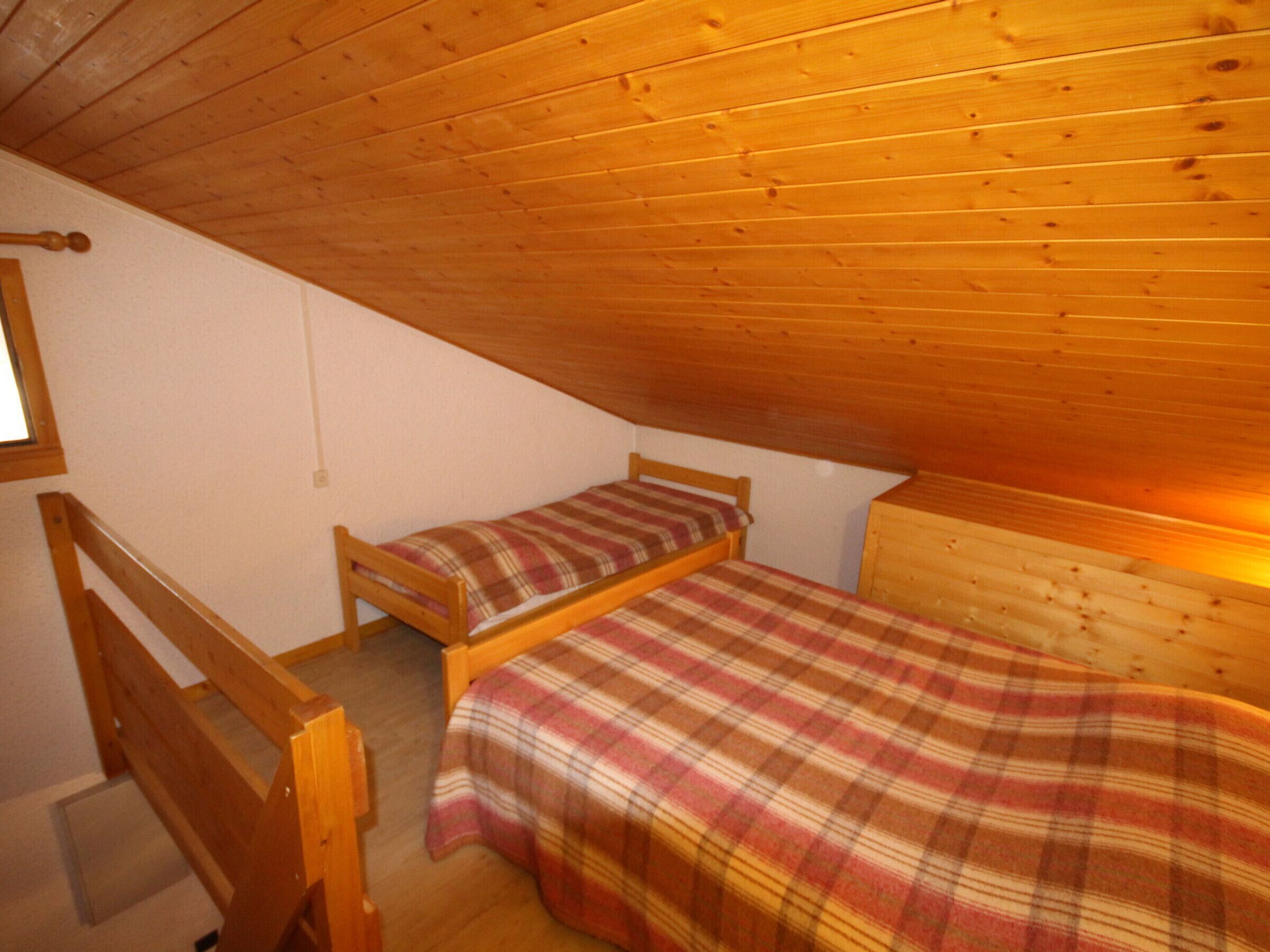 A small, cozy attic bedroom with wooden walls and a sloped ceiling. There are two wooden beds with plaid bedding.