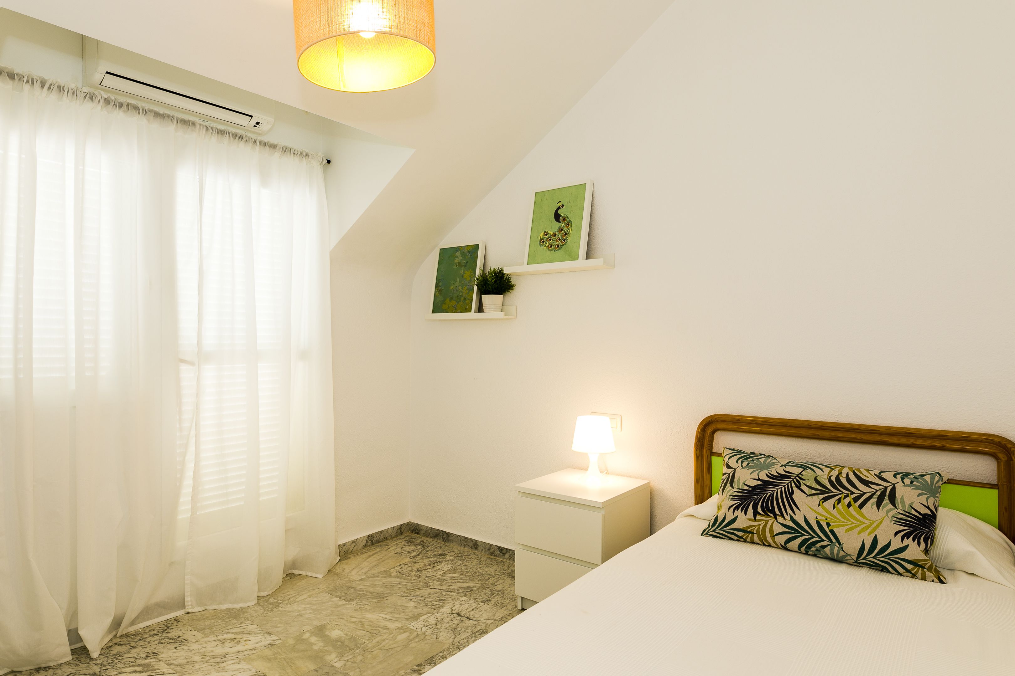 A bedroom with a bed, nightstand, wall-mounted shelf, and a window with sheer curtains.