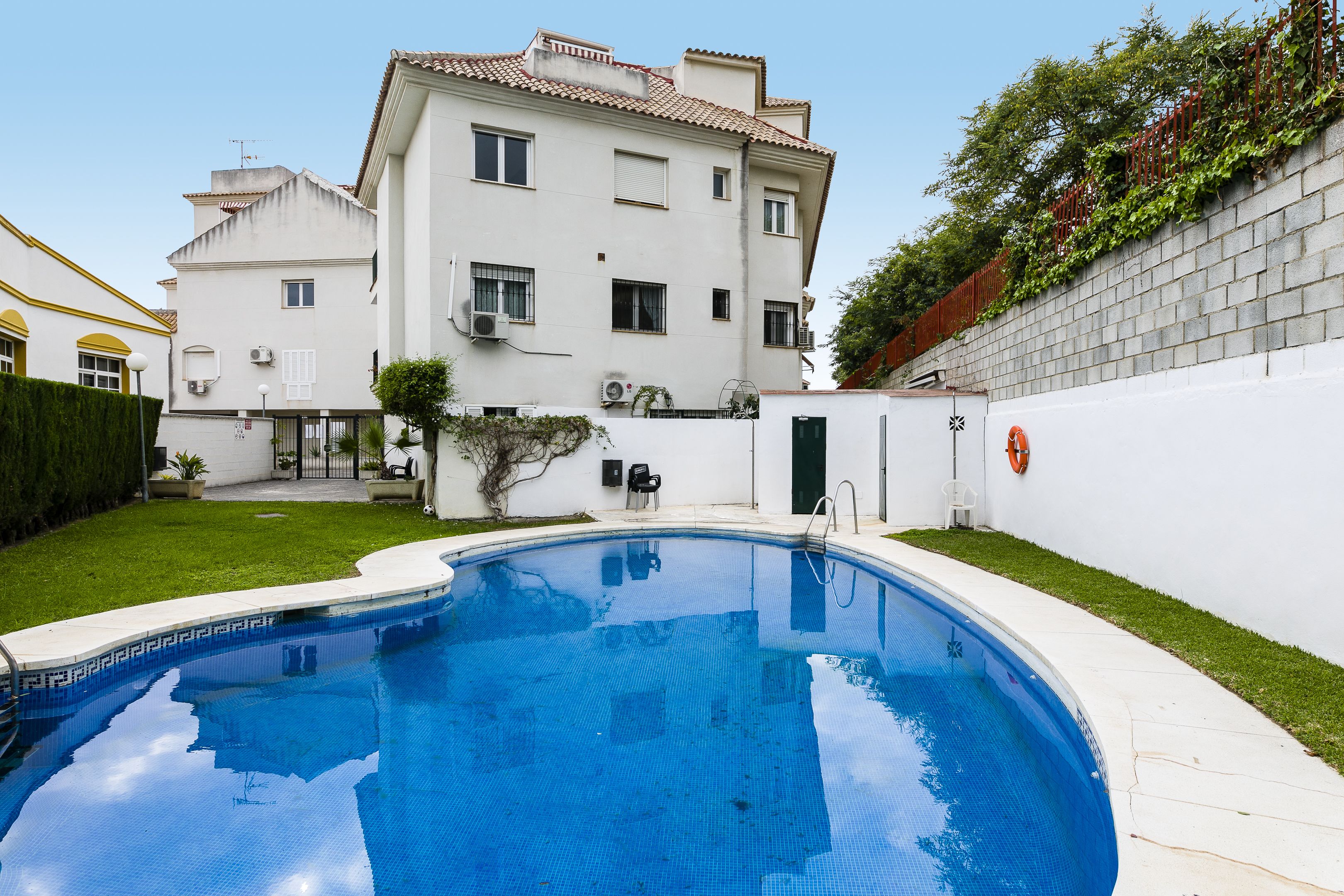 A swimming pool with a tiled deck, a white building with multiple windows and balconies, and a brick wall with greenery.