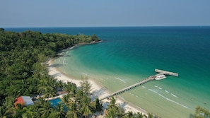 An aerial view of a beach with a pier extending into the ocean, surrounded by dense tropical vegetation.