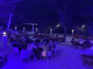 An outdoor dining area with tables and chairs, surrounded by palm trees and a thatched-roof structure.