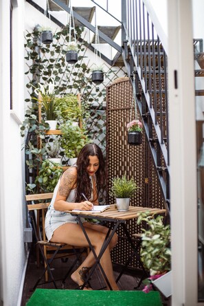 A woman sitting at a wooden table outdoors, writing. There are potted plants and a staircase in the background. - Deck Lodge - Hostel (Peniche)