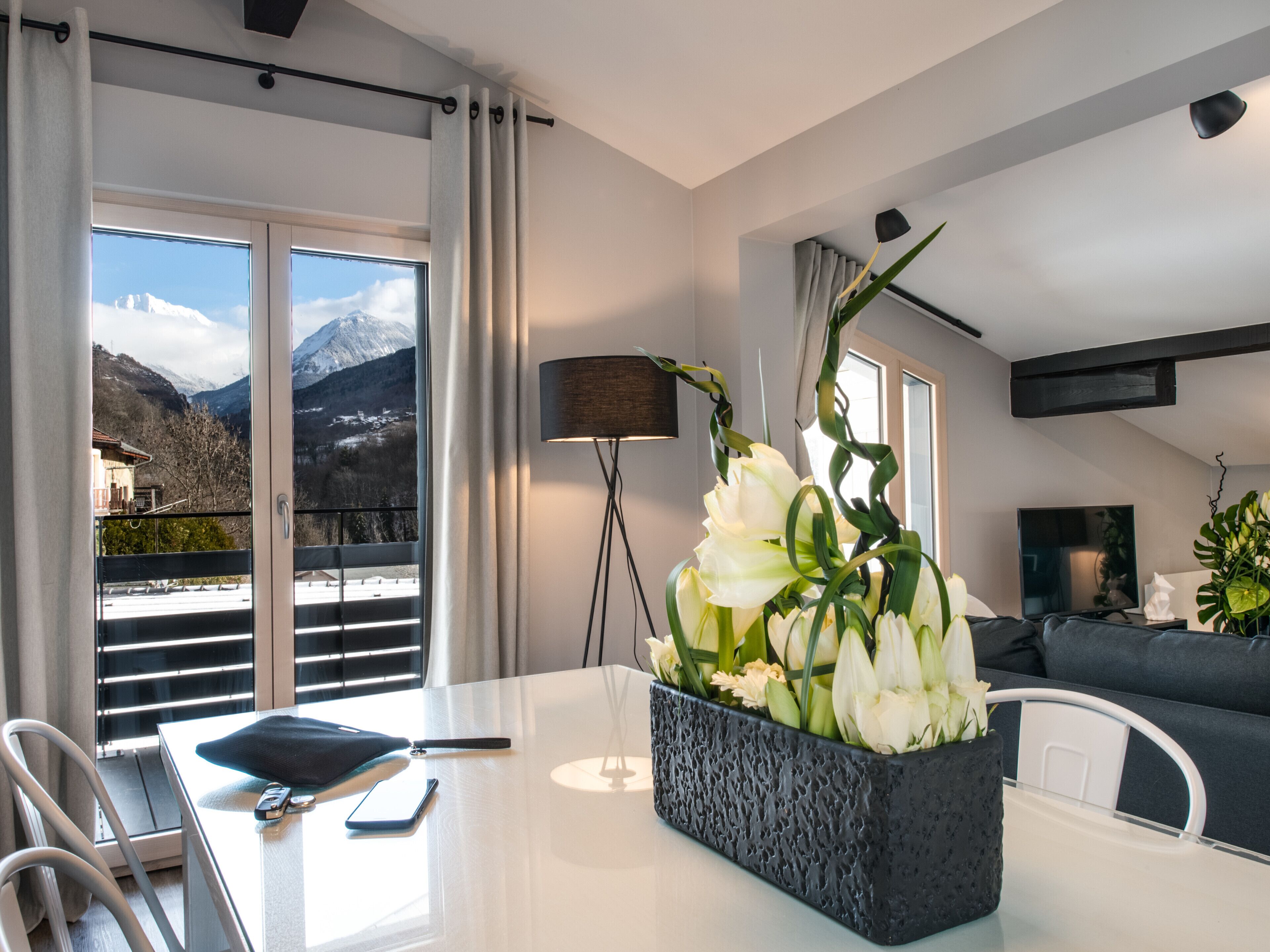 A modern hotel room with a dining table, chairs, a black vase with white flowers, a sofa, a TV, and a mountain view through large windows.