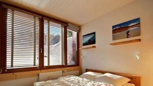 Un dormitori amb llit de fusta, grans finestres amb persianes i dos quadres al mur.