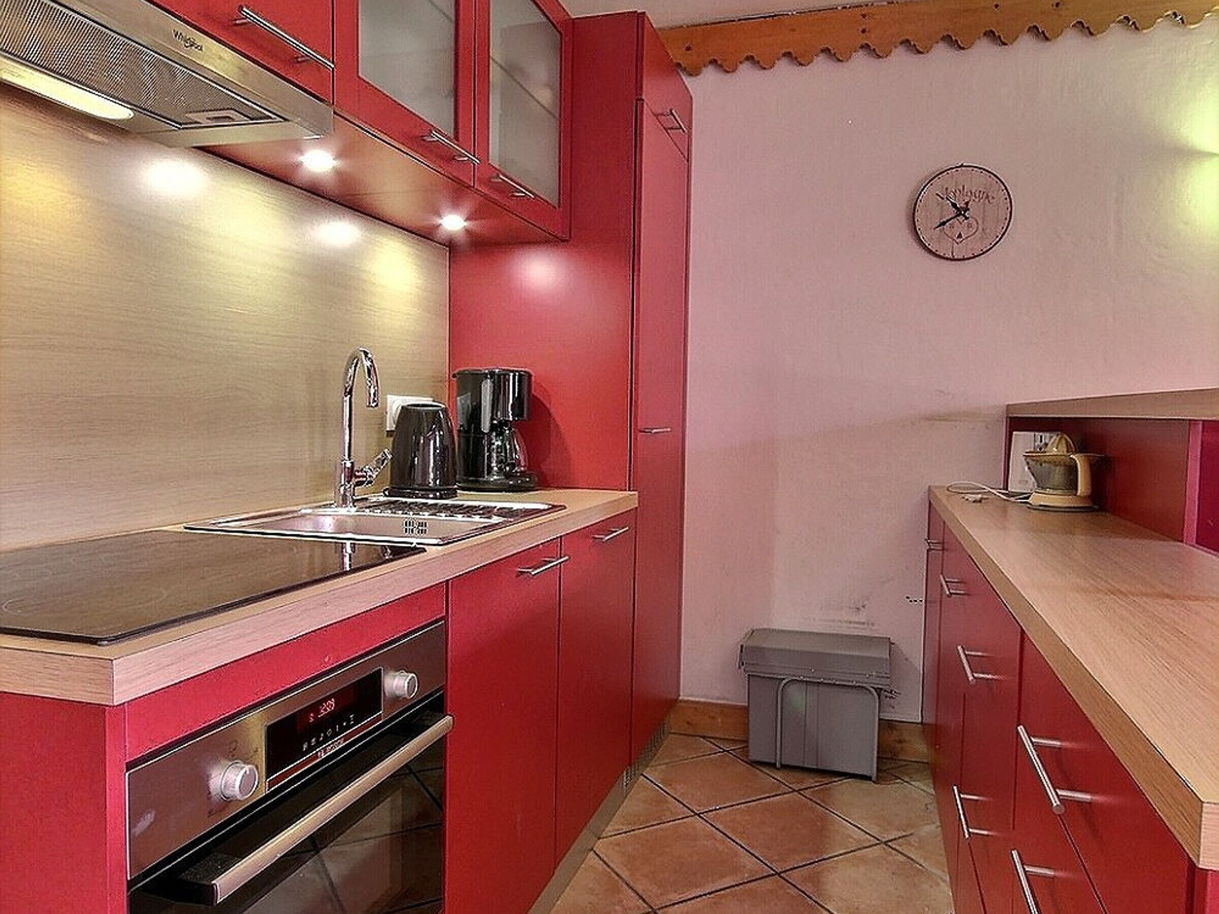 A compact kitchen with red cabinets, a stainless steel sink, a coffee maker, and a small trash bin.