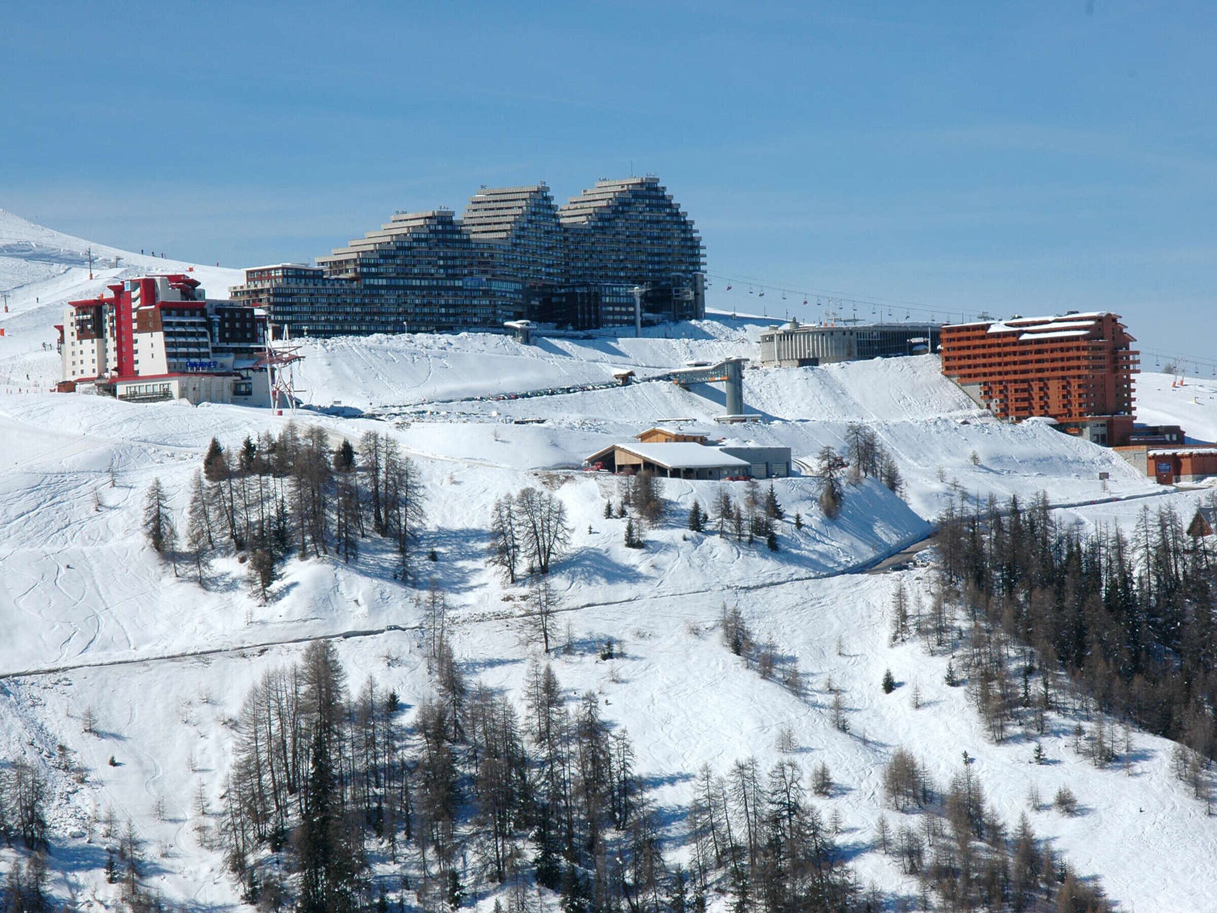 A ski resort with multiple buildings, including a large hotel with a red facade, and a ski lift in the background.