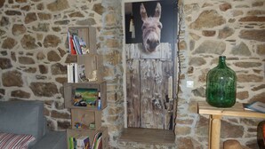 Una habitación rústica con pared de piedra, puerta de madera con cabeza de burro, sofá gris, un taburete pequeño y una mesa de madera con una botella verde.