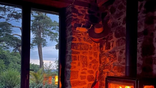 Una chimenea de piedra con fuego encendido, un banco rústico de madera y un ventanal con vistas a árboles y un camino de grava.