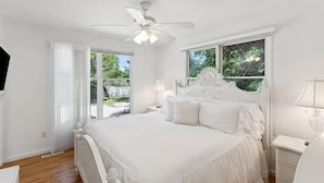 Une chambre à coucher blanche avec un grand lit, un ventilateur de plafond, une fenêtre donnant sur un paysage verdoyant et une table de chevet avec une lampe.