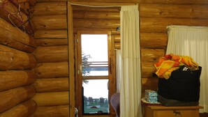 A log cabin interior with a wooden floor, a window with curtains, a wooden dresser, and a small table with items on it.