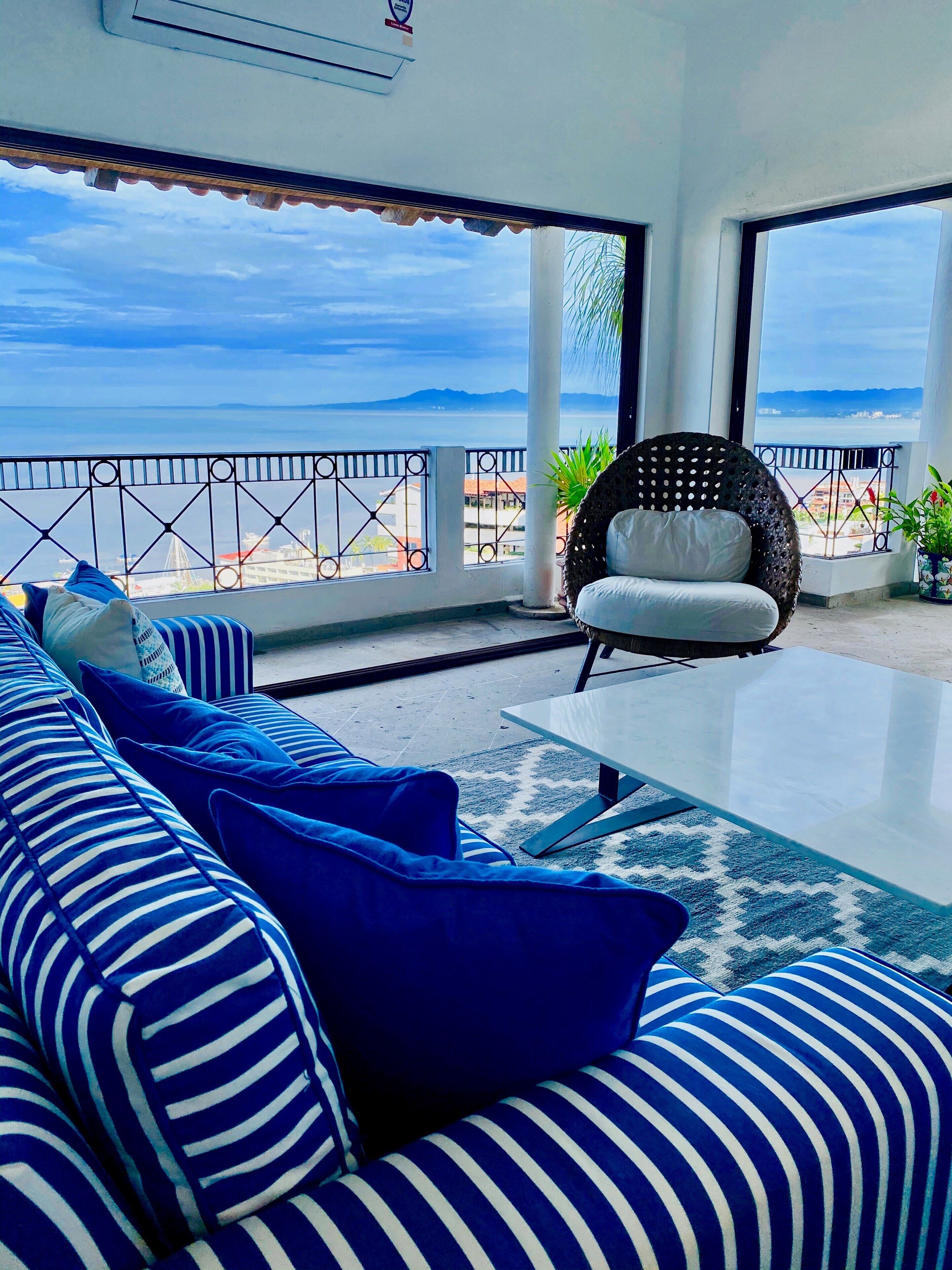 Un balcón con sofá y sillón, una mesa de vidrio y vista al mar.