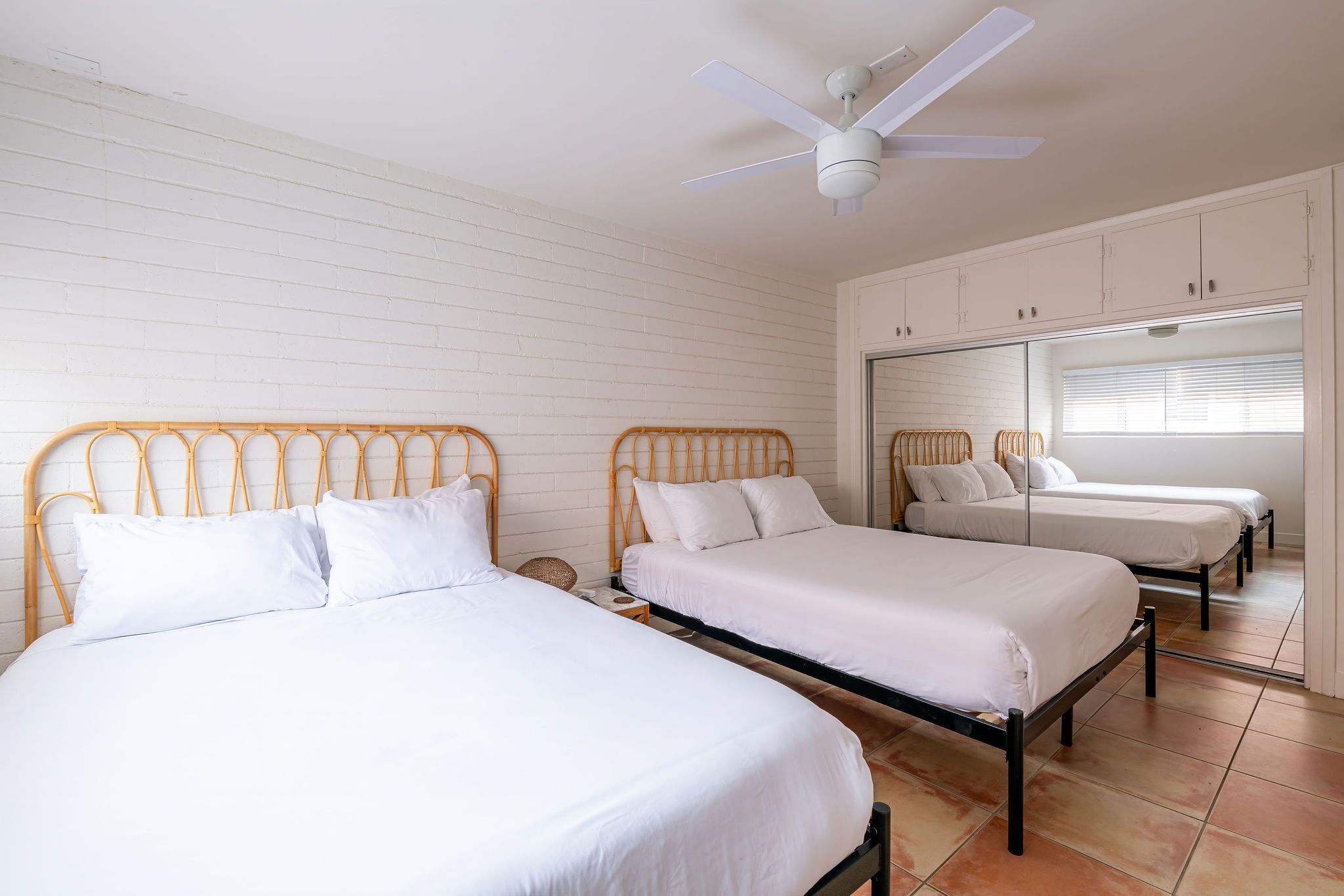 Une chambre avec deux lits simples, un ventilateur de plafond, des murs blancs et une fenêtre avec des stores.