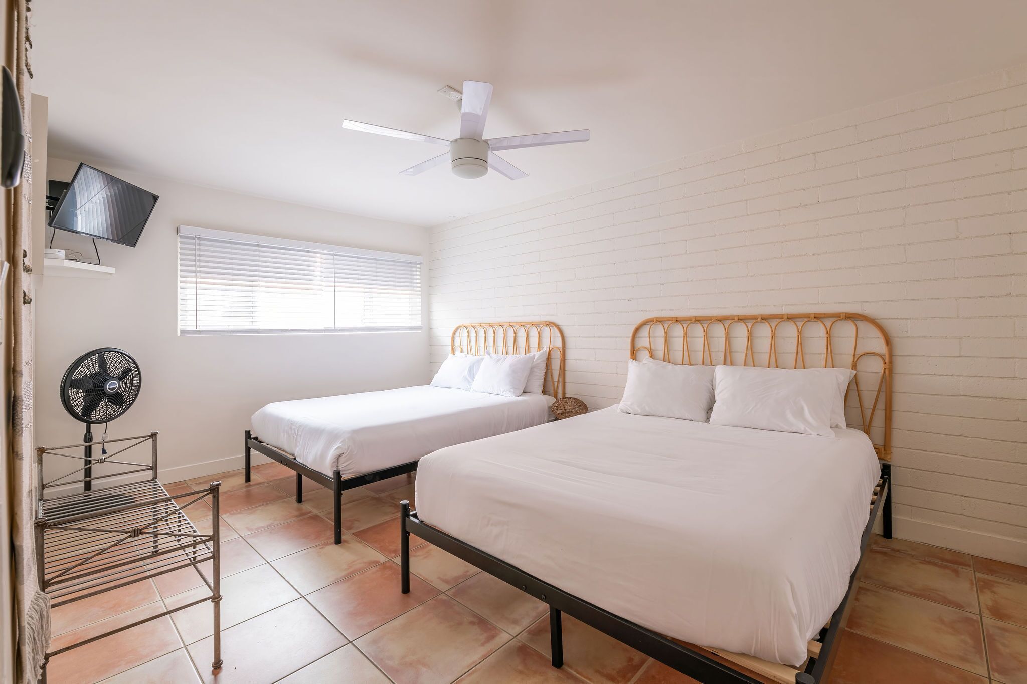 Une chambre avec deux lits, un ventilateur de plafond, un téléviseur posé sur une étagère et une fenêtre avec des stores.