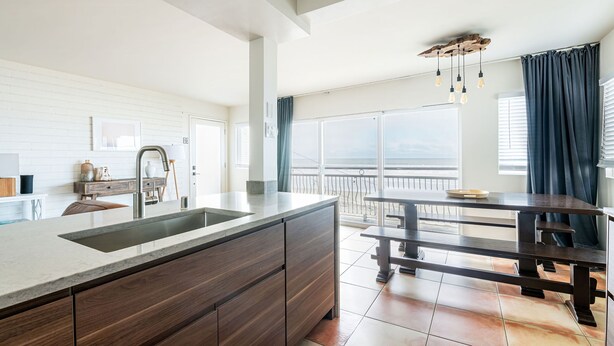 Une cuisine moderne dotée d’un grand îlot central, d’un coin repas avec un banc, et offrant une vue sur la mer grâce à de grandes fenêtres.