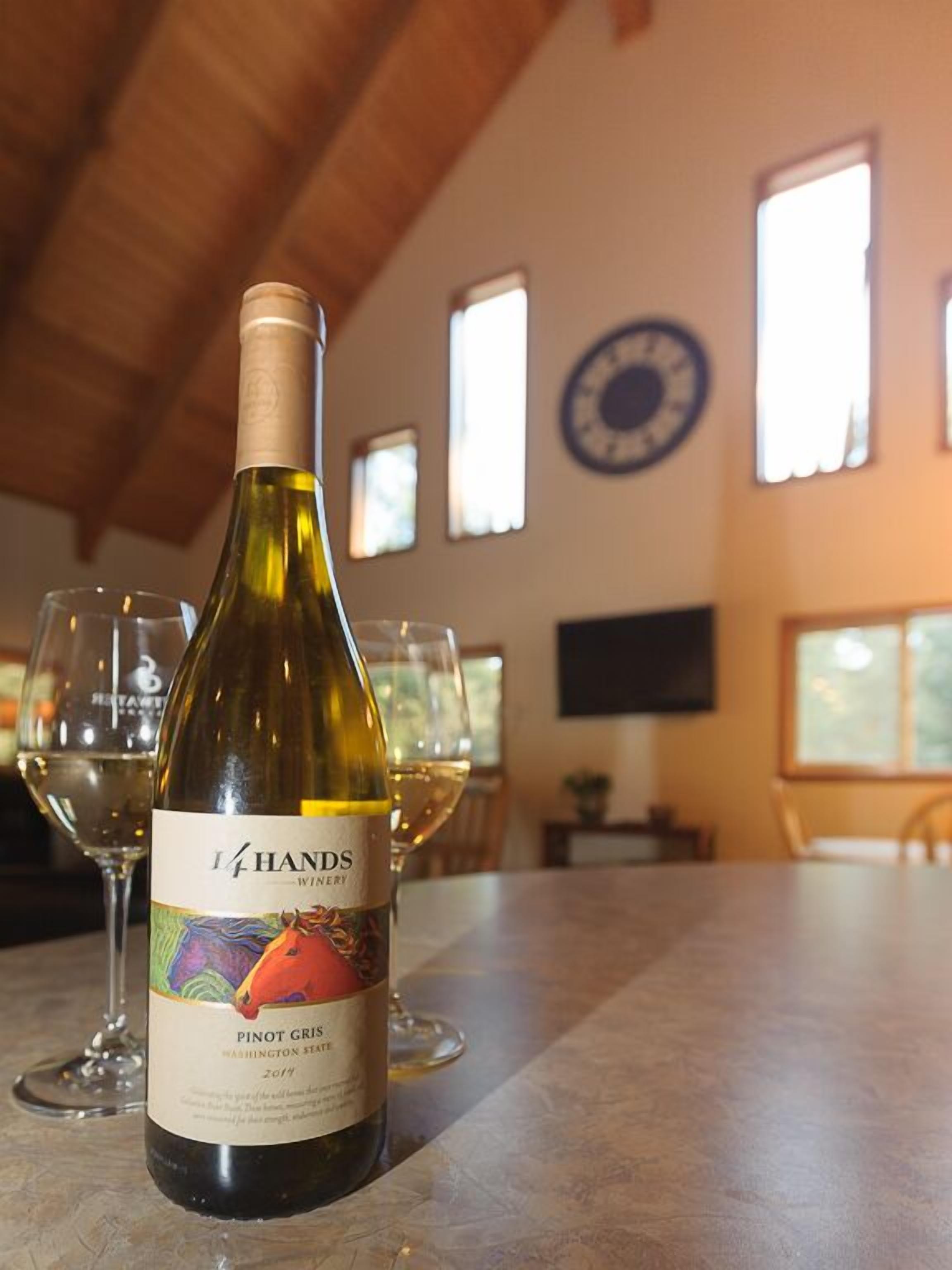 Une bouteille de vin est posée sur une table, entourée de verres, dans une pièce dotée de fenêtres et d’une horloge au mur.