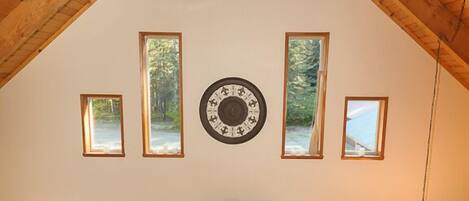 A cozy room with a high ceiling, wooden beams, a large clock, and multiple windows offering views of the outdoors.