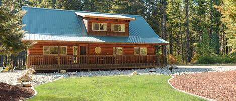 A log cabin with a blue roof, surrounded by a well-maintained lawn and a forested backdrop.