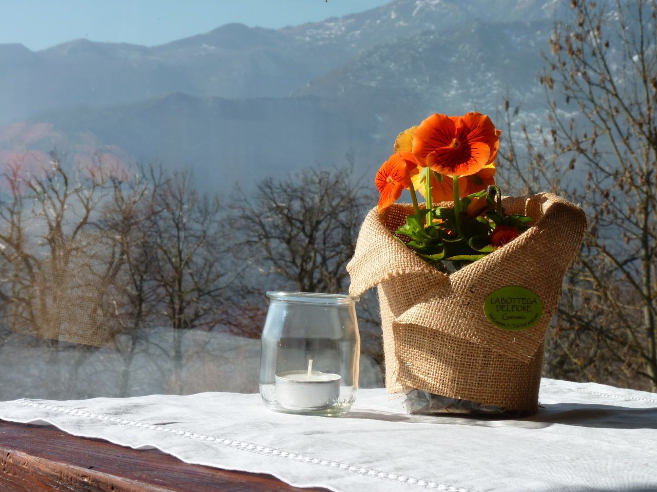 A candle in a glass jar, a bouquet of orange flowers in a burlap sack, and a tablecloth on a wooden table with a mountainous backdrop.