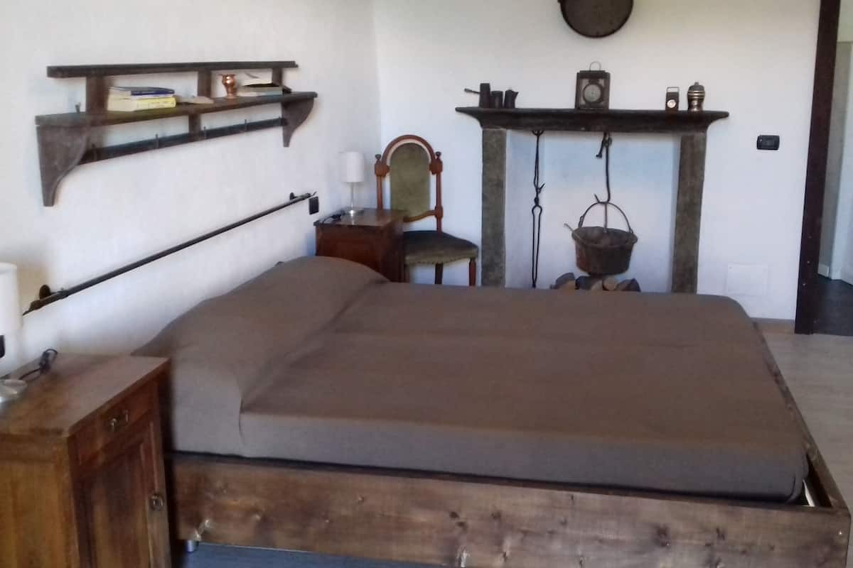 Una habitación con una cama de madera, una silla, una mesita con una lámpara, un estante con libros y una olla de cobre, y una chimenea con un reloj y una cesta colgante.