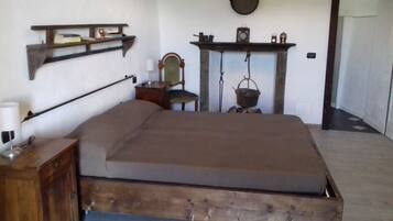 Een kamer met een houten bed, een stoel, een tafeltje met een lamp, een plank met boeken en een koperen pot, en een open haard met een klok en een hangmand.