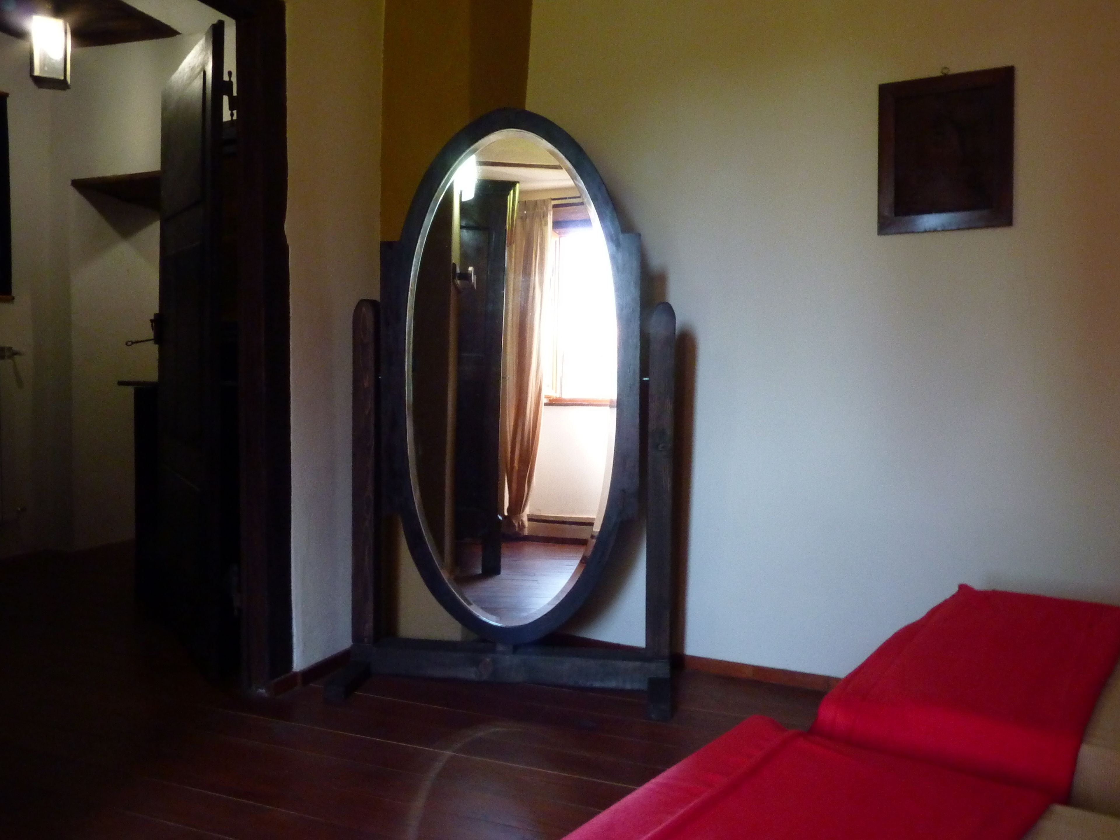 A room with a large oval mirror, a red sofa, wooden flooring, and a door leading to another room.