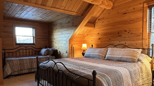 A cozy wooden cabin bedroom with a lofted sleeping area, a bed with a metal frame, and a nightstand with a lamp.