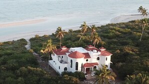 Um edifício branco com telhados vermelhos, rodeado por vegetação exuberante e palmeiras, próximo a uma praia.