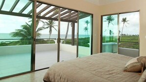 Quarto com uma cama grande, portas de vidro deslizantes que dão para uma varanda com vista para o oceano e palmeiras.