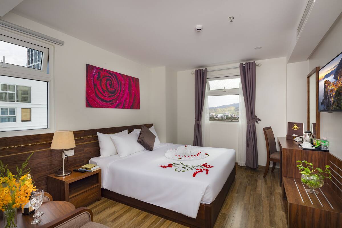 A hotel room with a large bed, a desk, a chair, a lamp, a vase with flowers, and a painting on the wall.