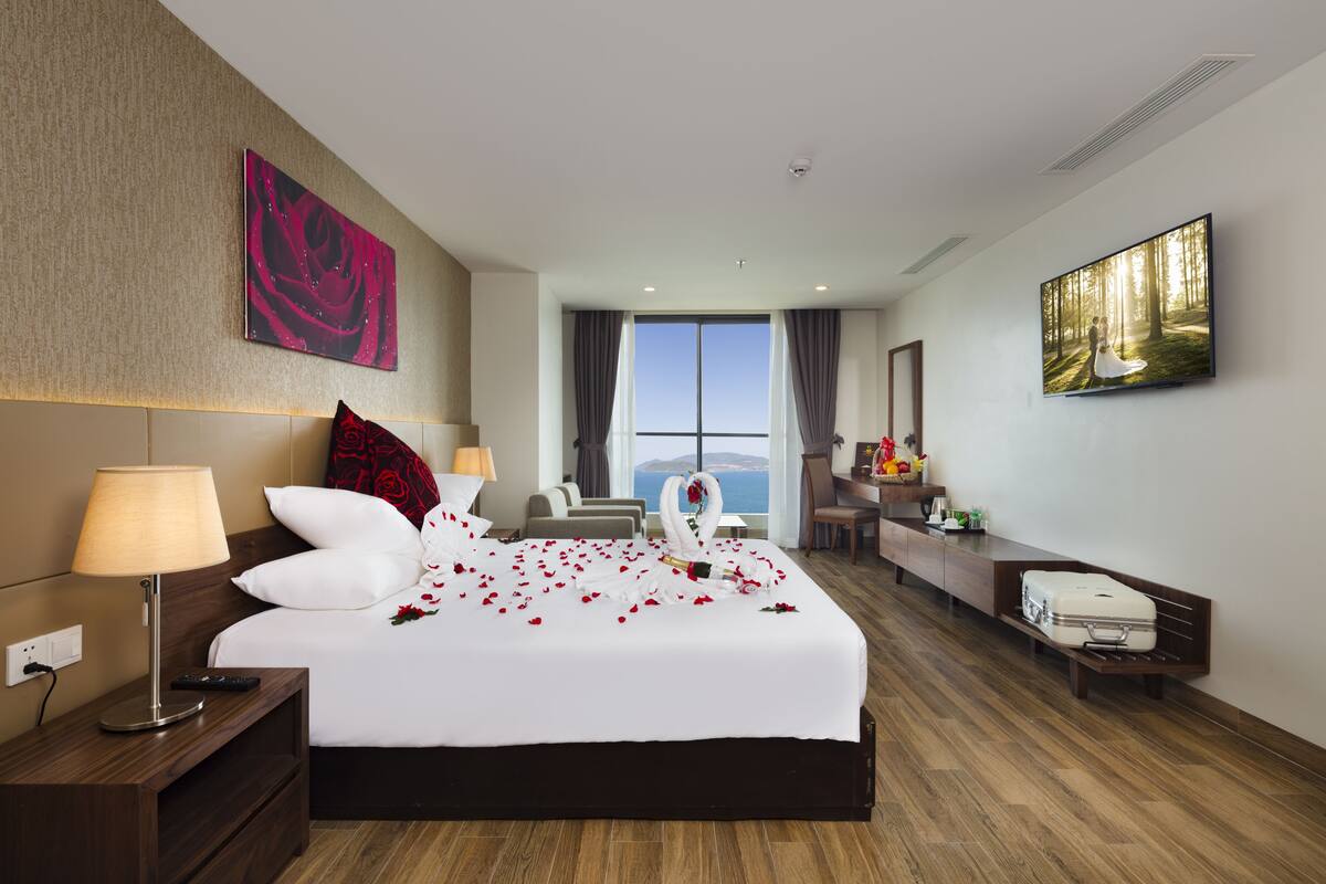 A hotel room with a large bed, a desk, a chair, a TV, and a view of the sea.