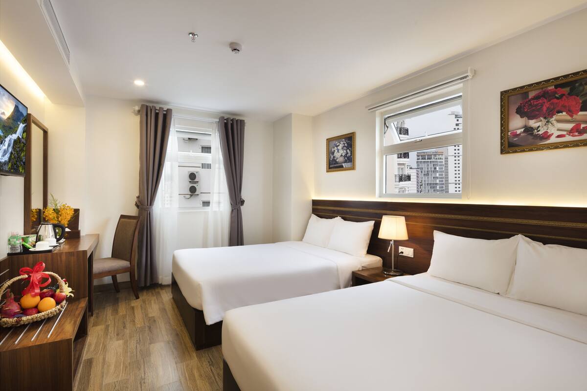 A hotel room with two beds, a desk with a basket of fruit, a chair, a window with curtains, and a framed picture on the wall.