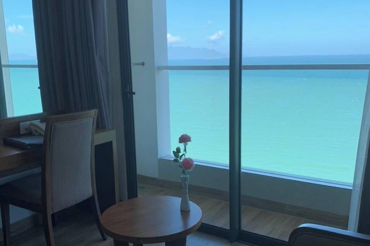 A hotel room with a large window offering a view of the sea. There is a wooden table with a vase of flowers, a chair, and a sofa.