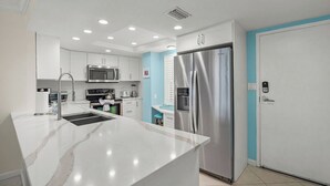 Cozinha moderna com armários brancos, geladeira de aço inoxidável, micro-ondas e pia com bancada de mármore.